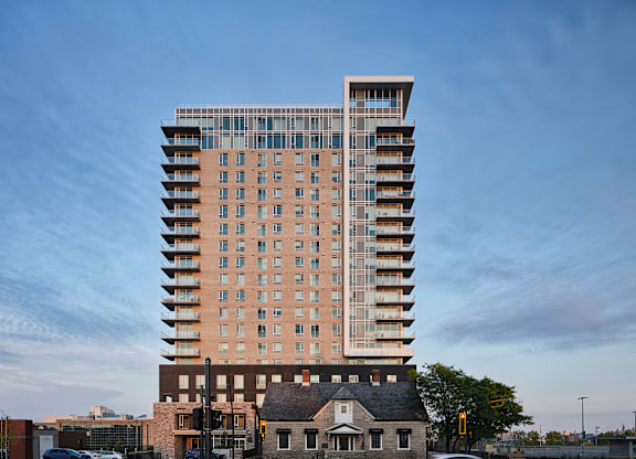 A tall building with a mix of brick and glass facade stands in front of a smaller house and a tree. at Le ColumbiaApartments , Gatineau, J8Y 0A8