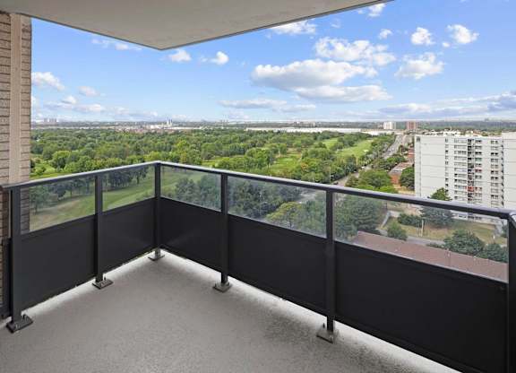 A balcony with a black railing overlooks a green landscape and buildings.