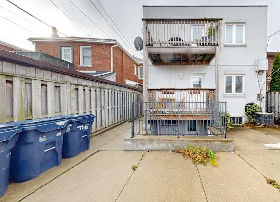 A grey building with a balcony is in the middle of a courtyard with blue bins and a fence.