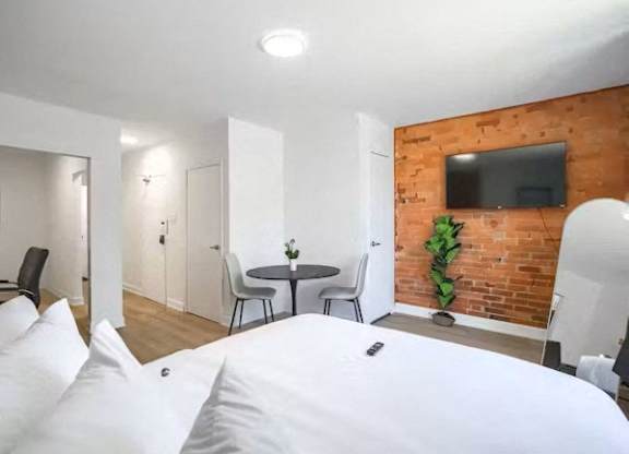 A bedroom with a bed, a table with chairs, and a brick wall with a TV.
