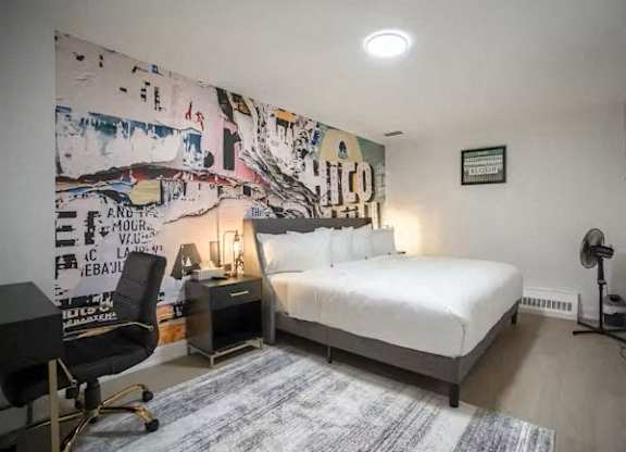 A bedroom with a bed, desk, chair, and a wall with a large artwork.
