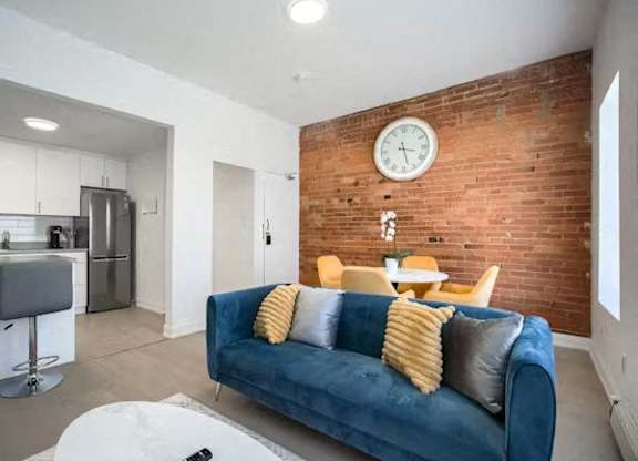 A blue couch with yellow pillows sits in a living room with a brick wall and a clock above it.
