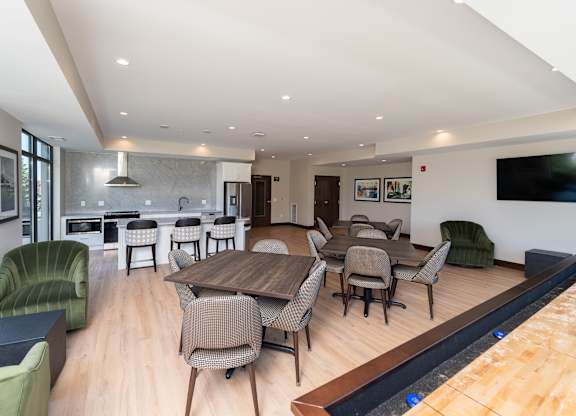 A modern community room kitchen with a dining table, comfortable chairs and shuffle board game.