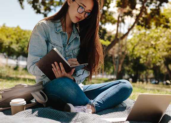 A person is sitting on a blanket outdoors, reading a book and using a laptop.