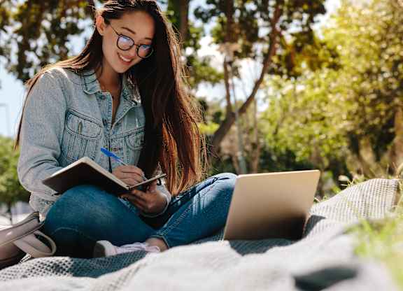 A person is sitting outdoors with a laptop and a notebook.