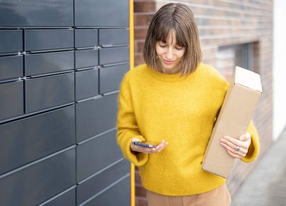 A woman in a yellow sweater is holding a box and a phone.