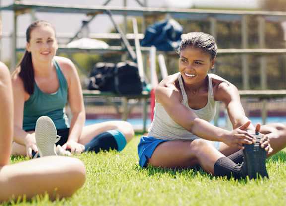 Three women are sitting on the grass, one of them is tying her shoelaces.