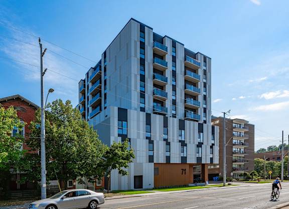 A modern multi-story building with blue and white balconies is situated on a street corner with a car and a cyclist in the foreground.