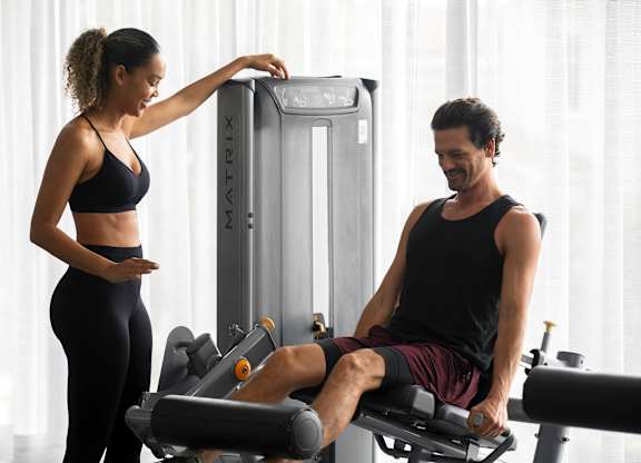 A woman is adjusting the settings on a gym machine while a man sits on a rowing machine. at Palma, Doral, FL