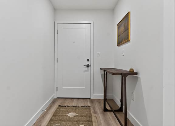 A hallway with a white door, a brown rug, and a brown table with a painting on the wall.