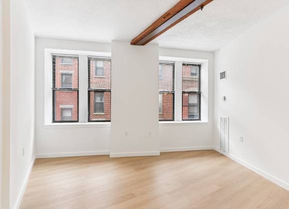 A room with wooden floors and two windows with red brick buildings outside.