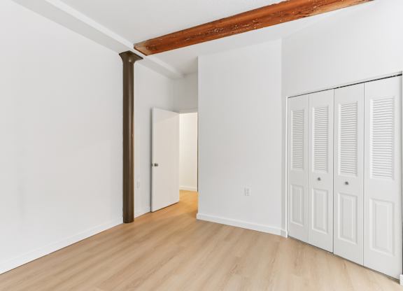 A room with white walls and wooden floors has a door on the left and three white closet doors on the right.