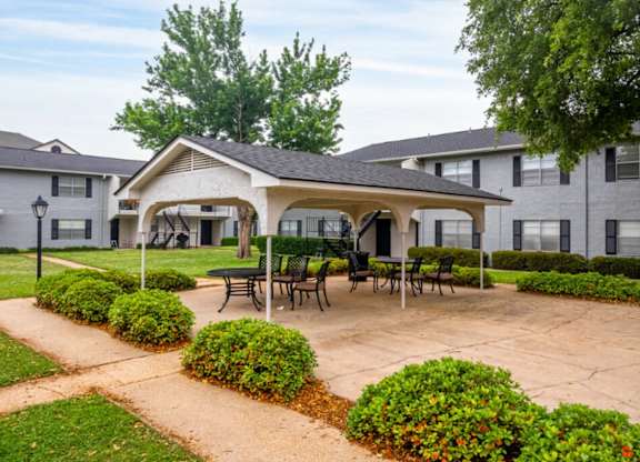 A patio with a table and chairs is surrounded by bushes and apartment buildings.