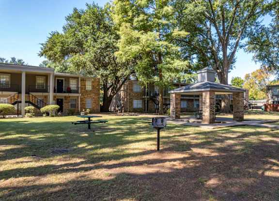 A small gazebo sits in the middle of a grassy area with apartment buildings in the background.