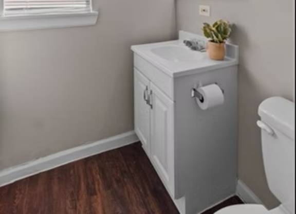 A bathroom with a white cabinet, a mirror with lights, a plant, and a window with blinds.