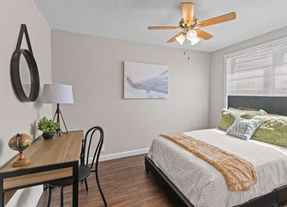 A bedroom with a bed, a table, a chair, a ceiling fan, and a painting on the wall.