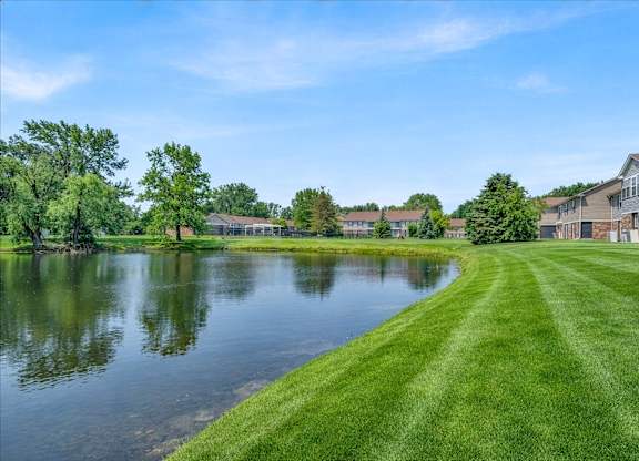A serene landscape with a lake, grass, and residential buildings.