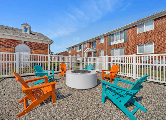 A white fence surrounds a fire pit with orange and blue chairs in front of a brick building.