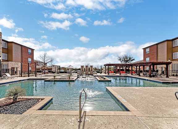 A pool area with a fountain in the middle and buildings in the background.