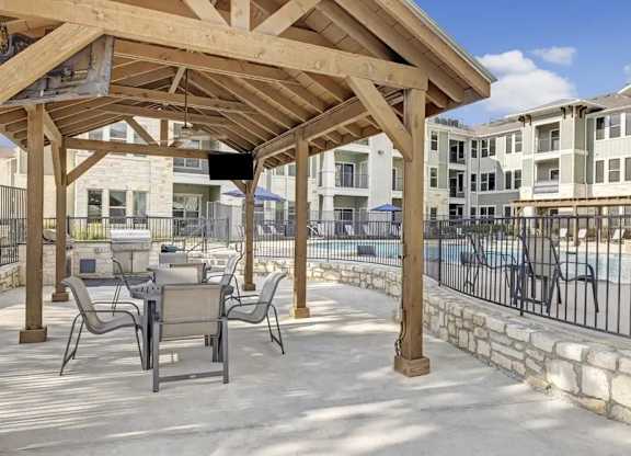A wooden pergola is over a patio with chairs and a pool in the background.