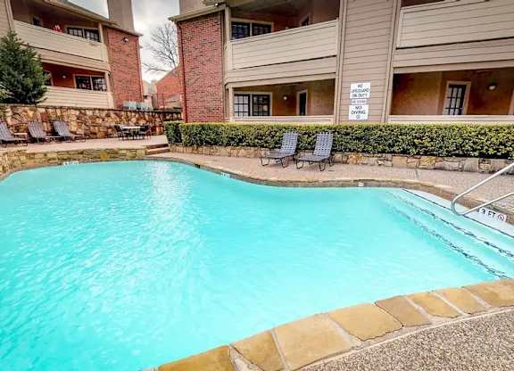 A swimming pool in front of a building with a stone wall and lounge chairs.