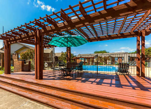 A wooden pergola over a pool with chairs underneath.
