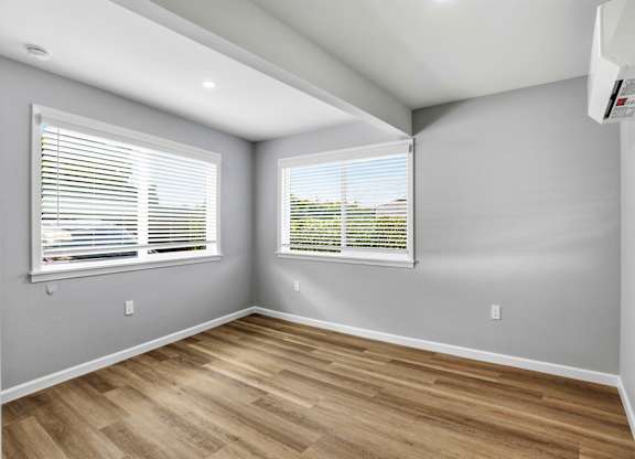 A room with wooden flooring and two windows with blinds.