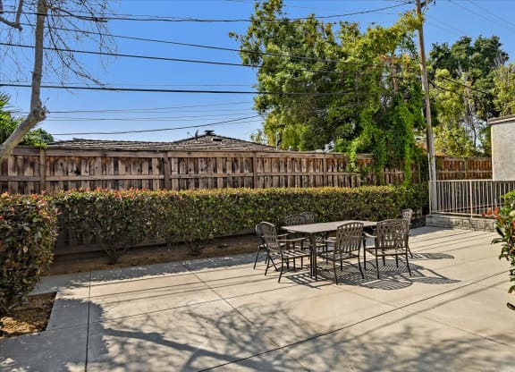 A patio with a table and chairs is surrounded by a hedge and a wooden fence.