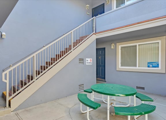A green table and chairs are on a patio outside a building with a staircase.