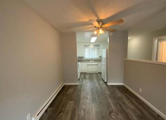 A room with a ceiling fan and a kitchen in the background.