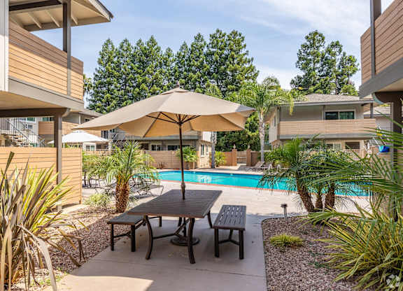 A patio with a table and chairs is surrounded by plants and a pool.