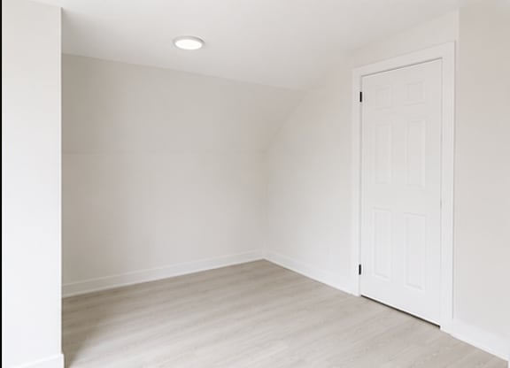 A white door is on the right side of a room with white walls and a light wooden floor.