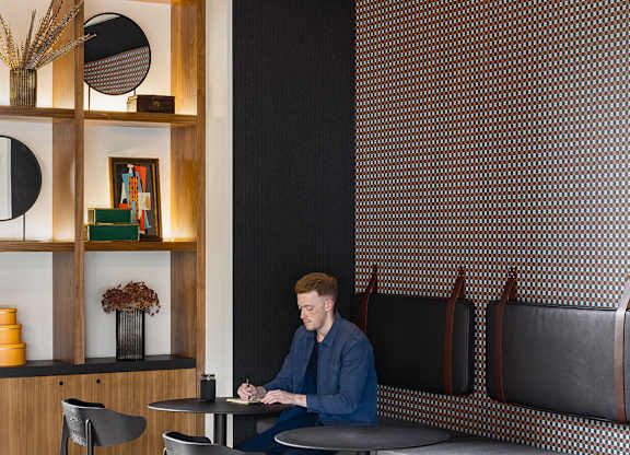A man is sitting at a table in a room with a wooden shelf and a black and white patterned wall.