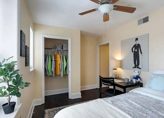 A bedroom with a bed, a ceiling fan, a chair, a table, a potted plant, and a painting of a person.