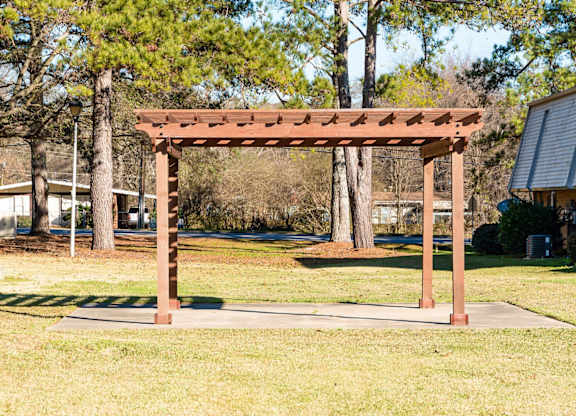 A wooden pergola stands in a grassy area with trees in the background.