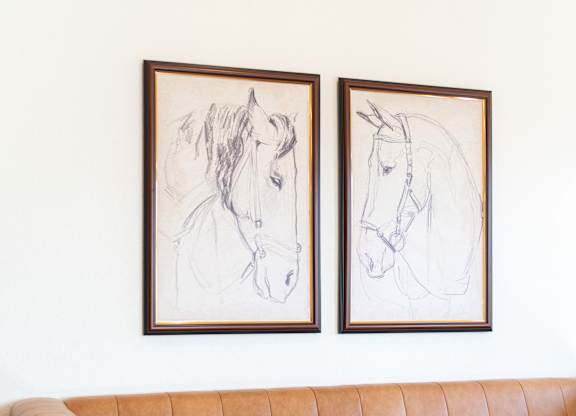 Two framed horse drawings hang on a wall above a tan couch.