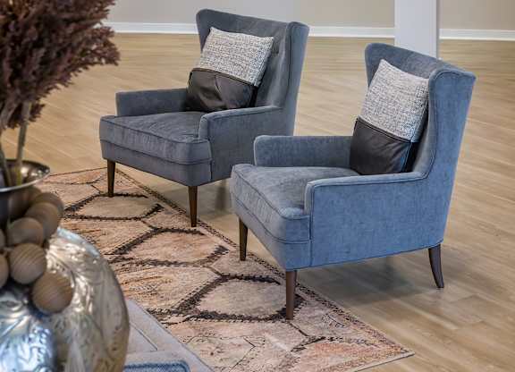Two grey armchairs with pillows are placed in a room with a large vase and a rug.