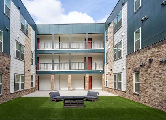 A courtyard with a grass lawn and a bench in front of a building with a balcony.