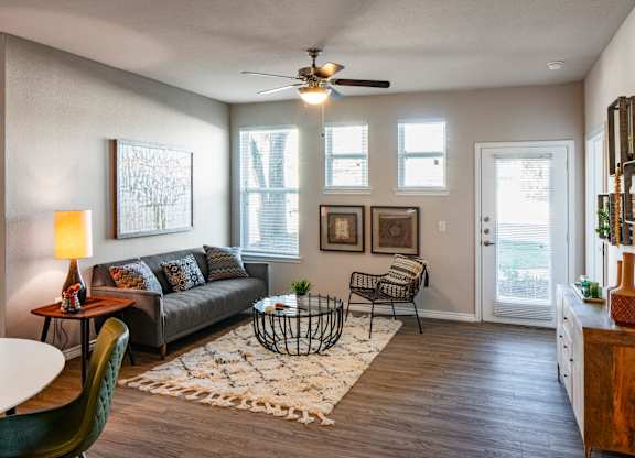 A living room with a grey couch, a green chair, a white table, a rug, a ceiling fan, and a door leading to a balcony.