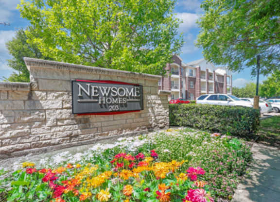 A sign for Newsome Homes stands in front of a building and flowers.