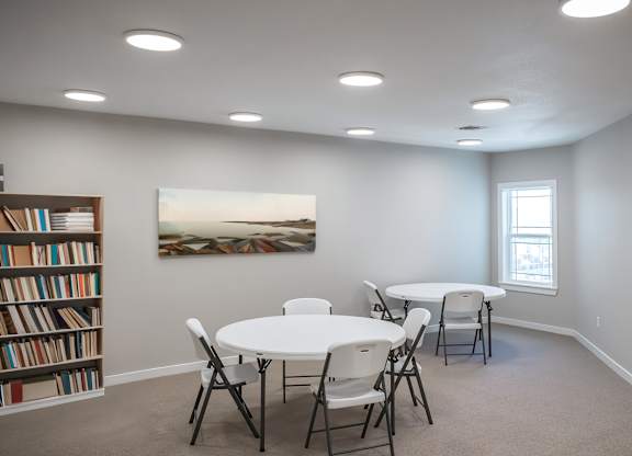 A room with a white table and chairs, a bookshelf, a clock, and a painting on the wall.