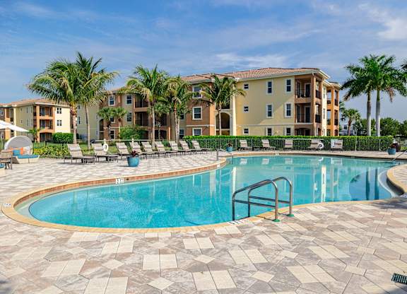 A swimming pool surrounded by a brick patio and palm trees in front of apartment buildings.