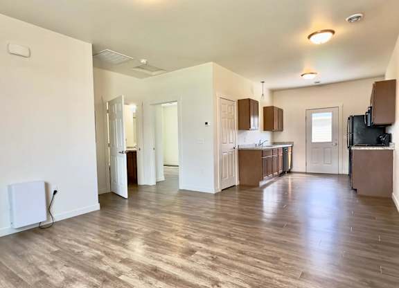 A spacious living room with wooden flooring and a kitchen area in the background.
