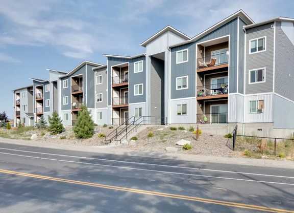 A row of modern apartment buildings with balconies and pine trees.