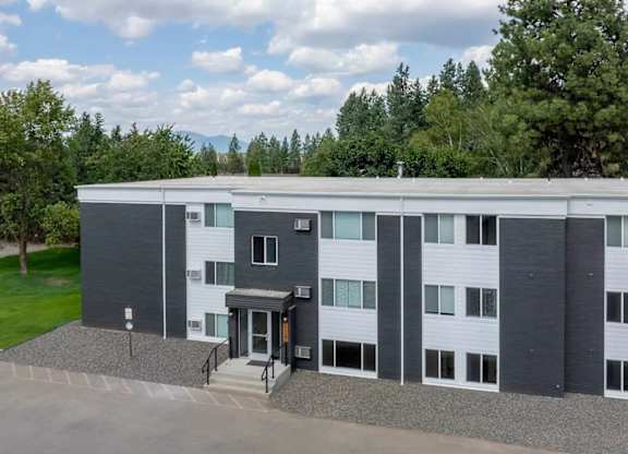 A modern building with a grey and white exterior is surrounded by trees and has a gravel driveway in front.