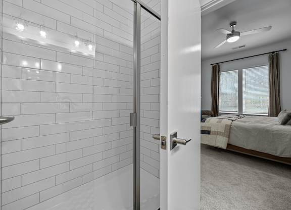 A white tiled bathroom with a white door and a bedroom with a bed and a ceiling fan.at Address East End, Raleigh North Carolina