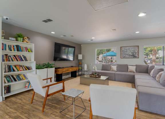 A living room with a grey couch, a white chair, a wooden chair, a white bookshelf with books, a television, and a window with a view of a blue awning and palm trees.