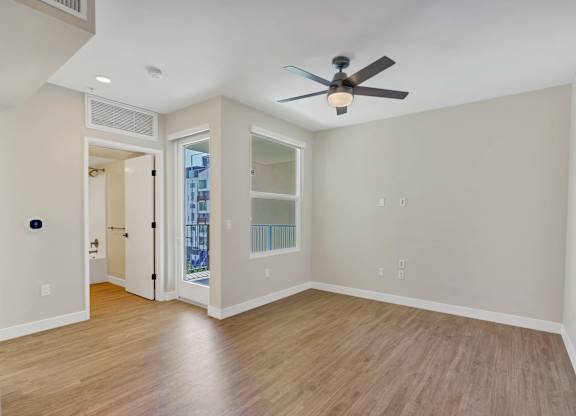 A room with a ceiling fan and light, hardwood floors, and a door leading to a balcony.