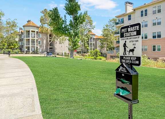 A sign for a pet waste station is on a pole in a grassy area.