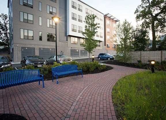 A brick walkway leads to a building with a blue bench on the left.
